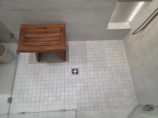 Walk-in shower with white square floor tile, a center drain and a teak shower bench