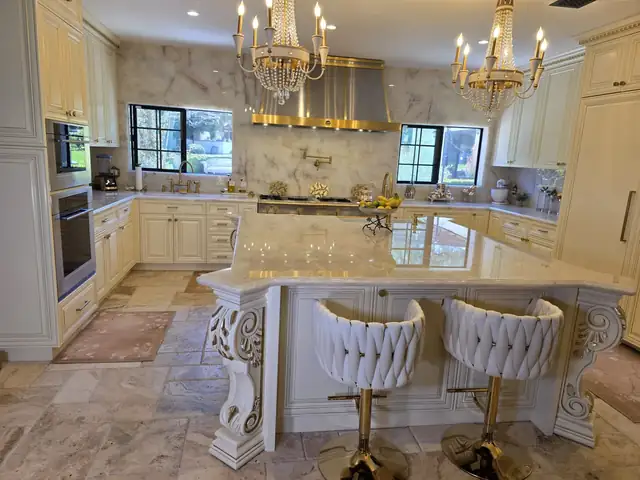 Kitchen by Better Remodels with cream cabinets, a marble island on carved legs, gold chandeliers and velvet counter stools